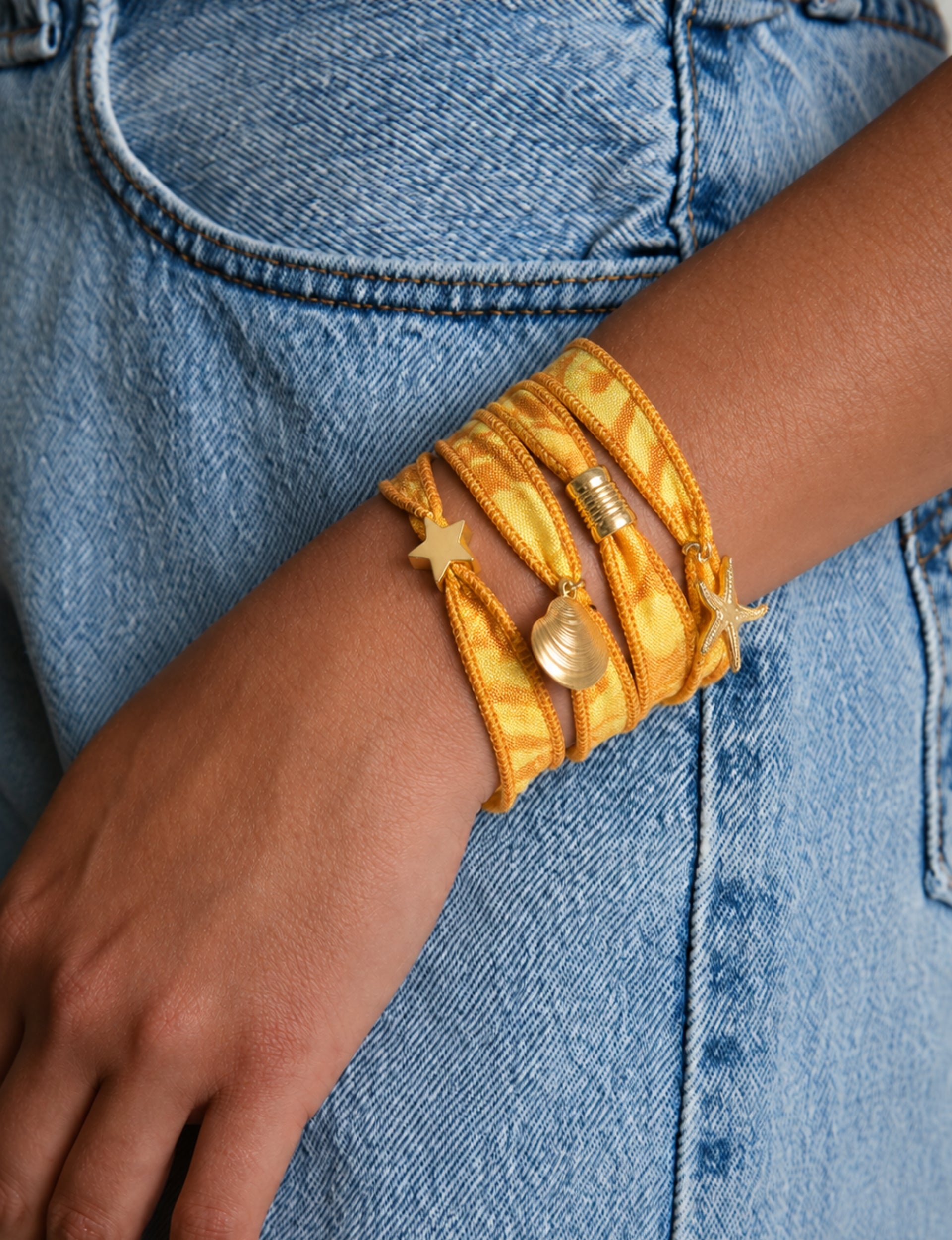 Orange abstract wrap-around fabric bracelet with gold star, shell, and starfish charms worn on wrist against blue denim jeans