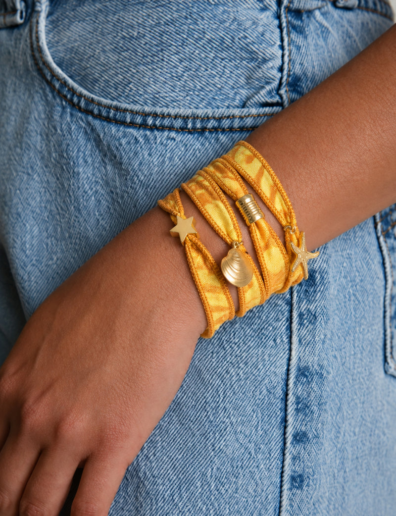 Orange abstract wrap-around fabric bracelet with gold star, shell, and starfish charms worn on wrist against blue denim jeans