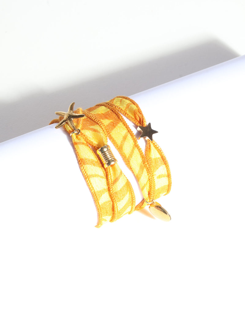 Orange abstract patterned wrap-around fabric bracelet with gold starfish, star, and shell charms, shown flat on white surface