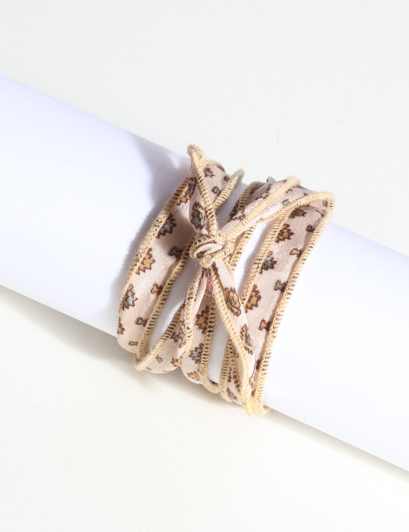 Beige fabric wrap-around bracelet with small brown and green abstract floral pattern and stitched edges, shown on white display