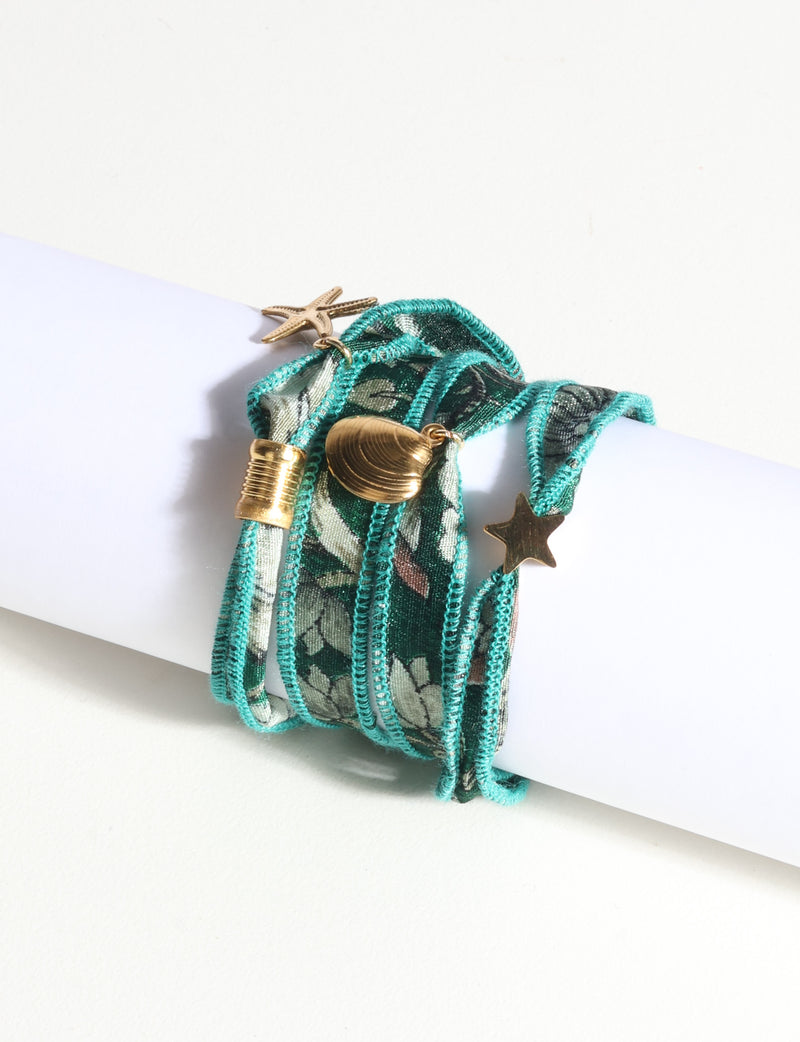 Turquoise wrap-around fabric bracelet with gold star, shell, and textured cylinder charms, shown on white display rod