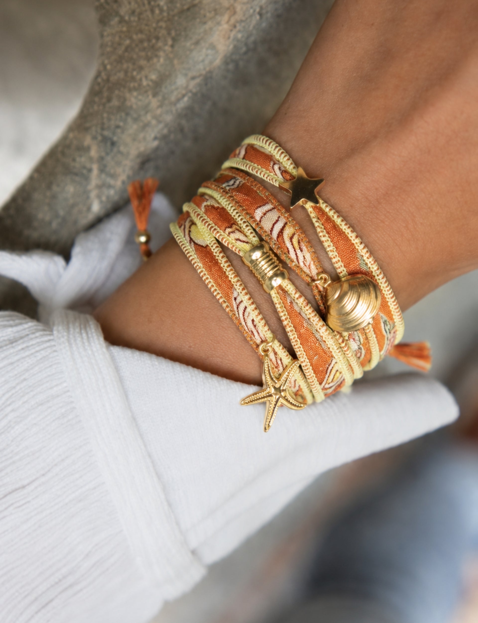 Orange and yellow wrap-around fabric bracelet with gold star, shell, and starfish charms worn on wrist