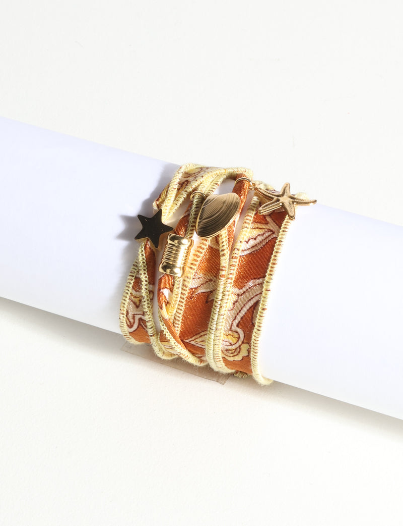 Orange abstract patterned wrap-around fabric bracelet with gold-tone star, shell, and charm embellishments, displayed flat