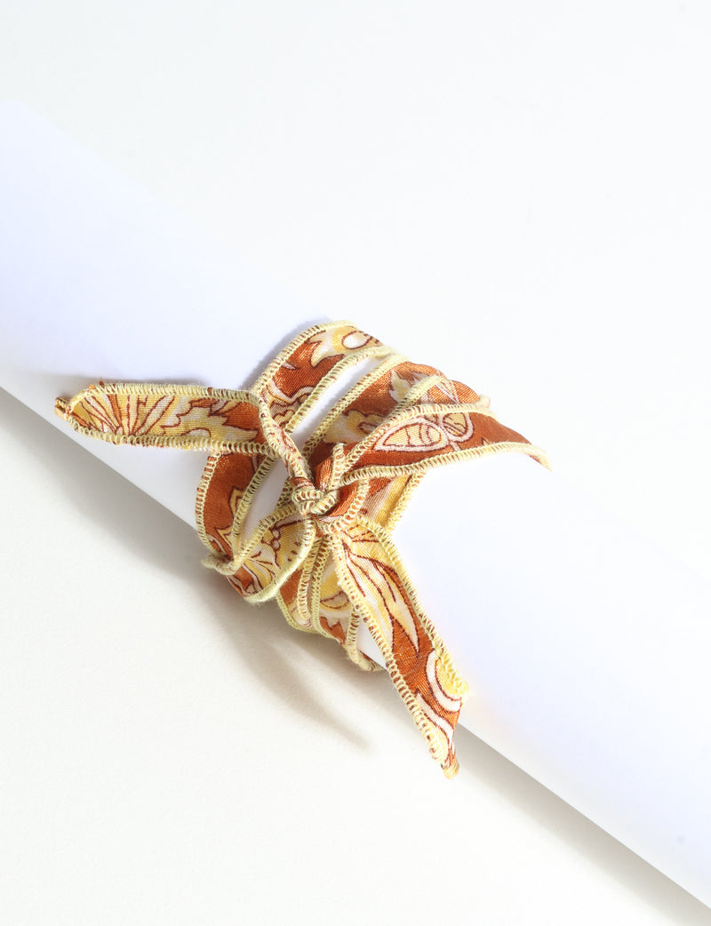 Orange abstract patterned fabric wrap-around bracelet with stitched edges and tied bow, shown flat on white background