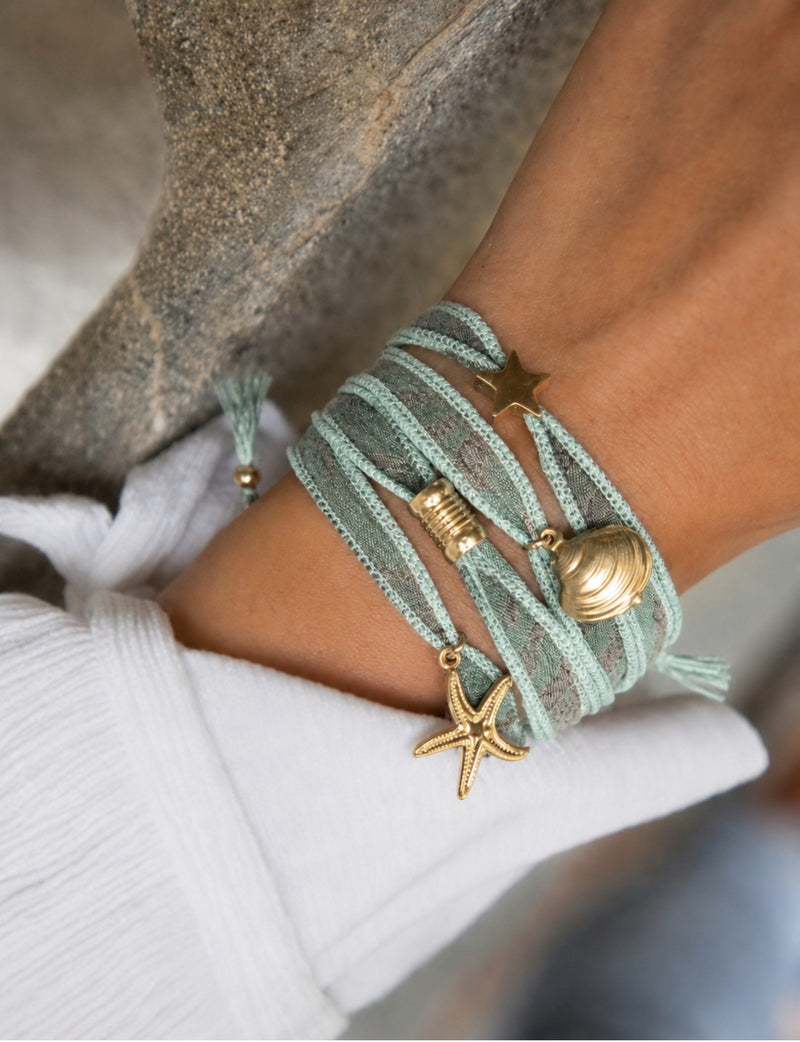 Light blue wrap-around bracelet with gold star, shell, and starfish charms worn on wrist with white sleeve visible