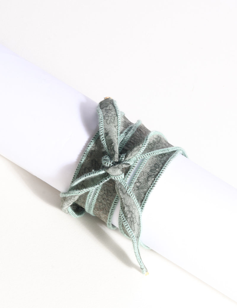 Green fabric wrap-around bracelet with stitched edges tied in a bow, shown on a white cylindrical display