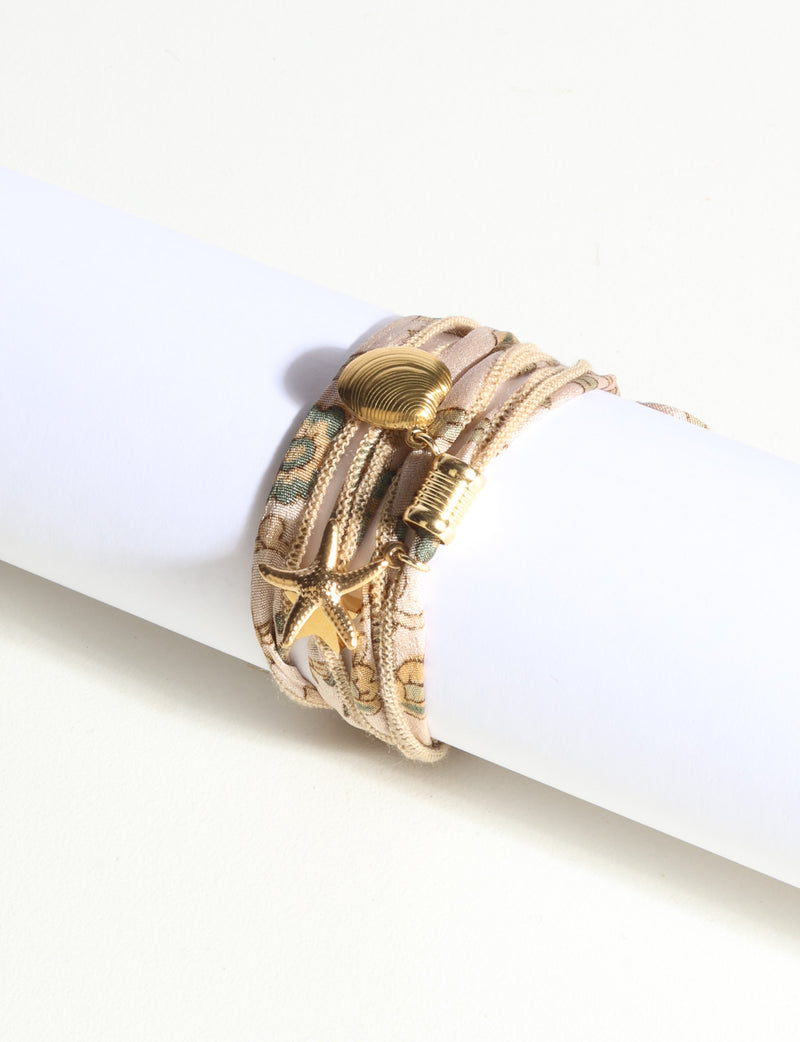 Wrap-around bracelet with beige and green abstract fabric, gold-tone shell, starfish, and barrel charms, displayed on white tube
