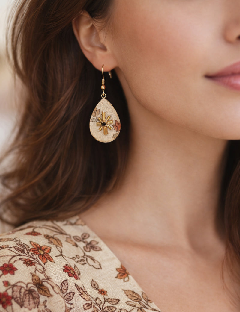 Beige teardrop earrings with tropical floral print and gold bead embellishments worn by a woman in a matching floral top