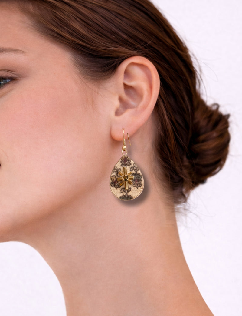 Beige teardrop earrings with tropical floral pattern and gold bead embellishment worn on a woman's ear, side view