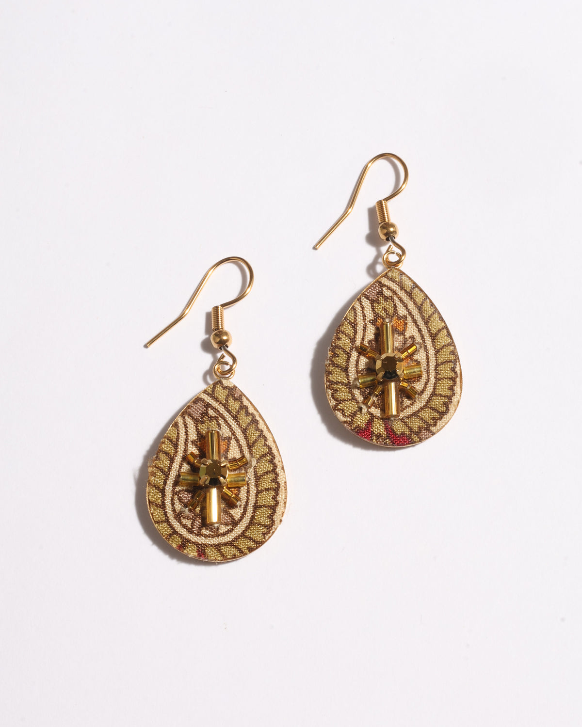 Beige tropical teardrop earrings with gold-tone hooks and bead embellishments on patterned fabric, flat lay view