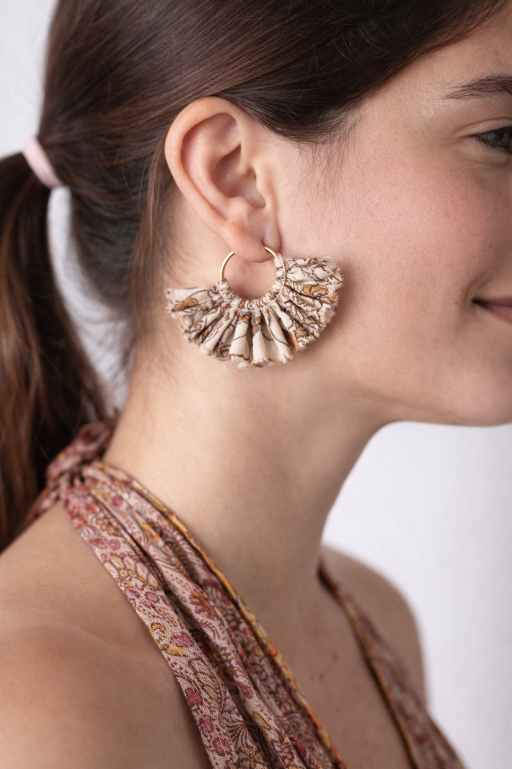 Beige tropical frill fabric hoop earrings worn by a woman with brown hair tied back and a patterned halter top