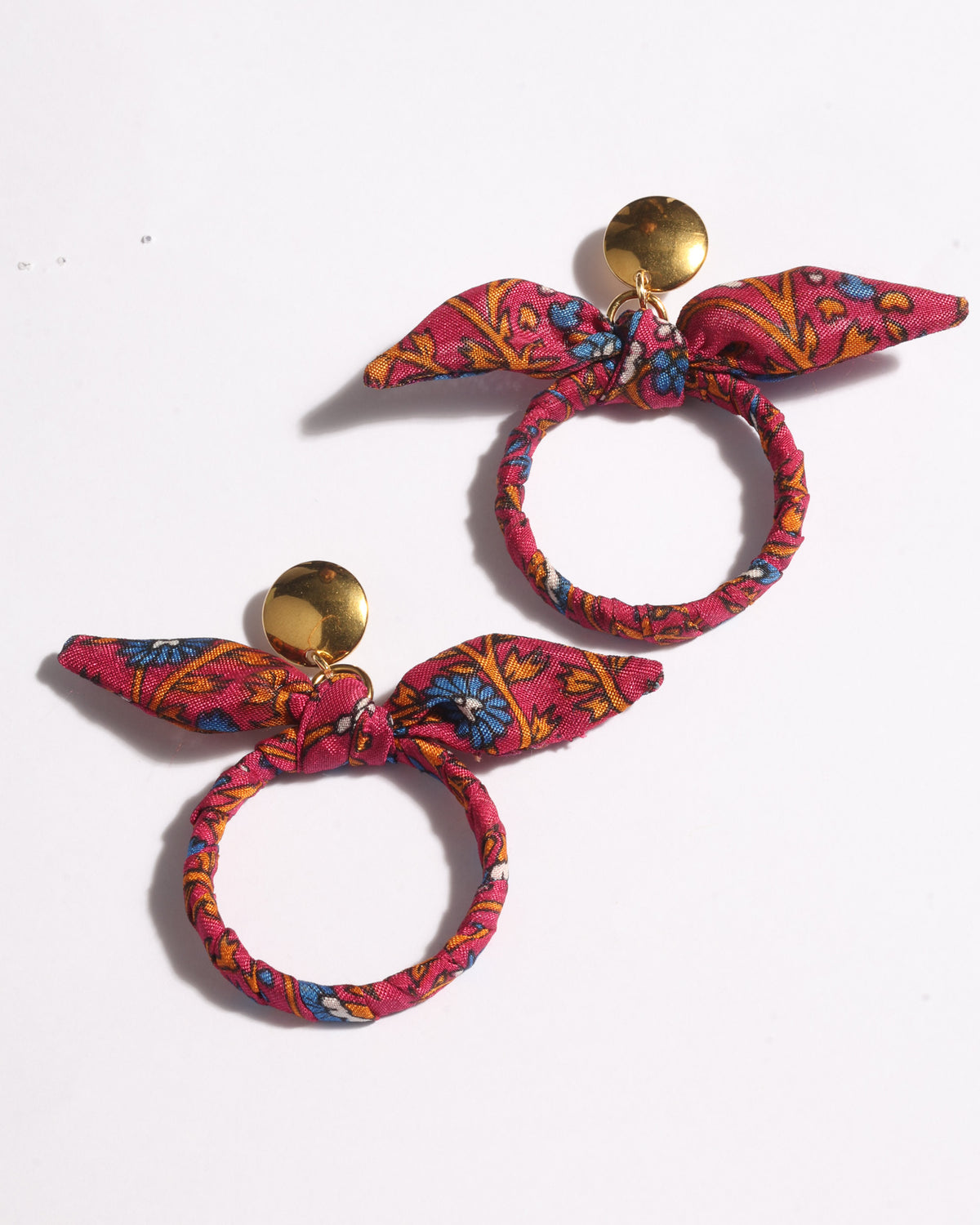 Pair of pink tropical bow earrings with gold circular studs and fabric-wrapped hoop detail, flat lay on white background