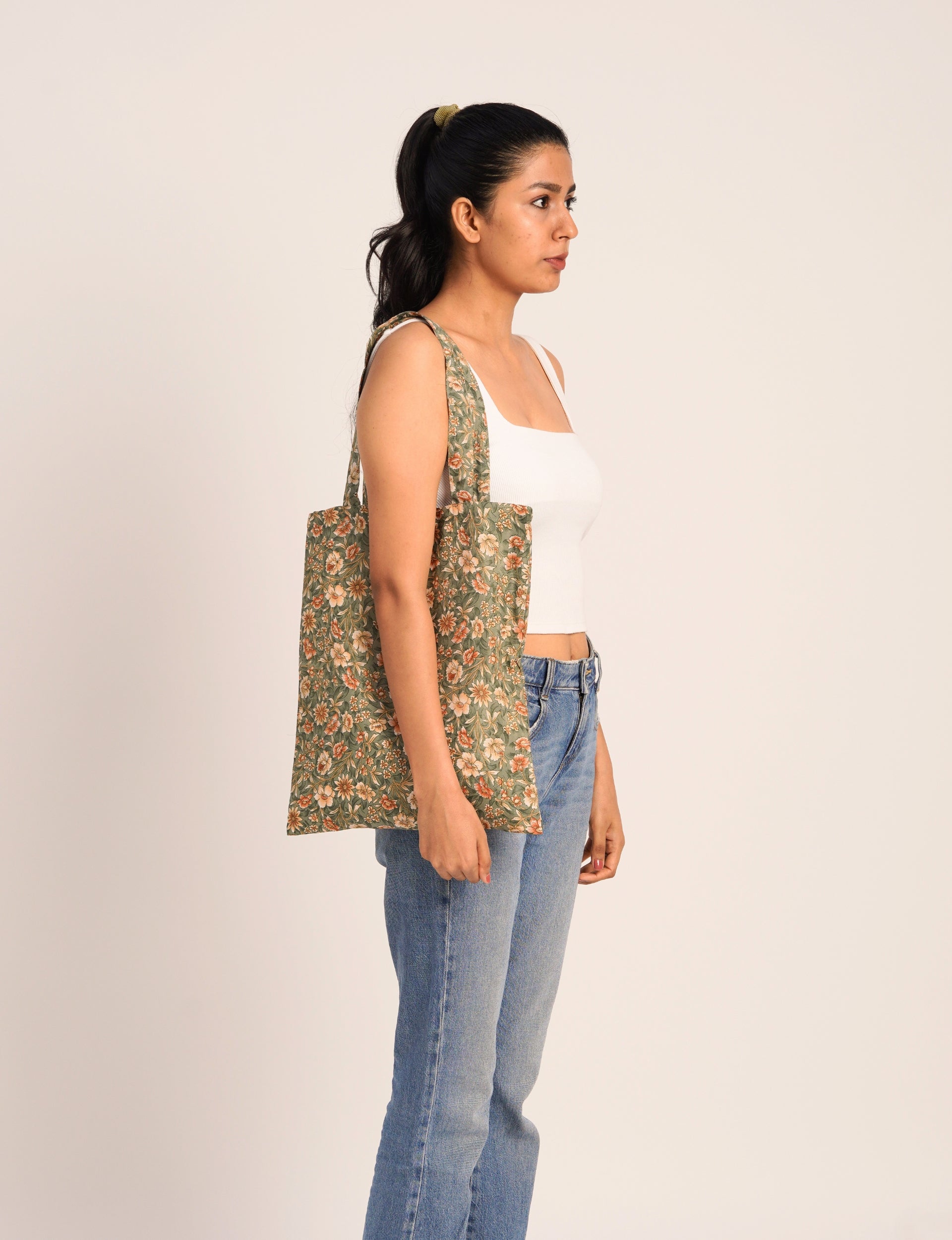 Woman wearing green floral print fabric tote bag over shoulder, shown in side view with white tank top and blue jeans