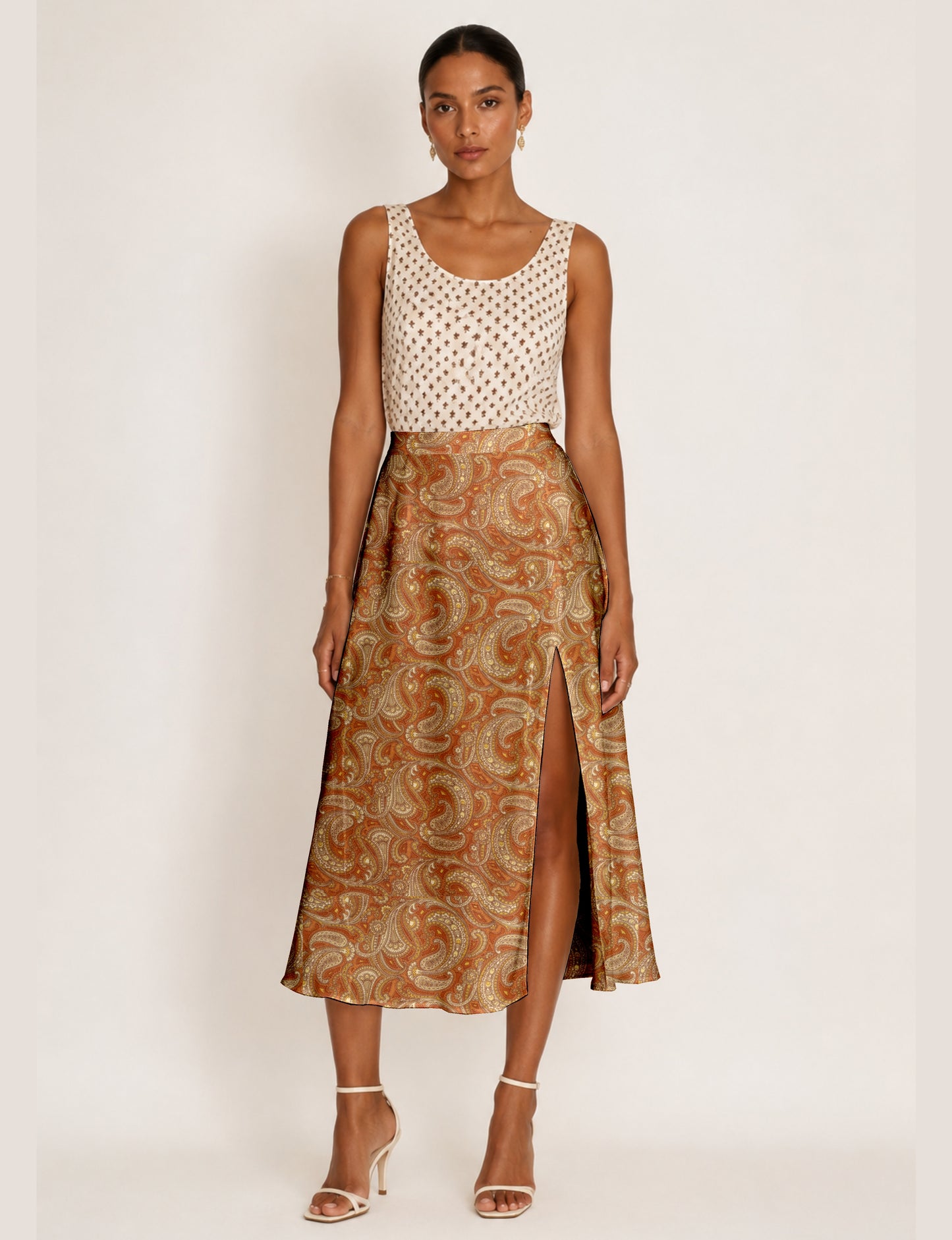 Woman wearing an orange paisley A-line skirt with a front slit, paired with a sleeveless white patterned top and beige heels