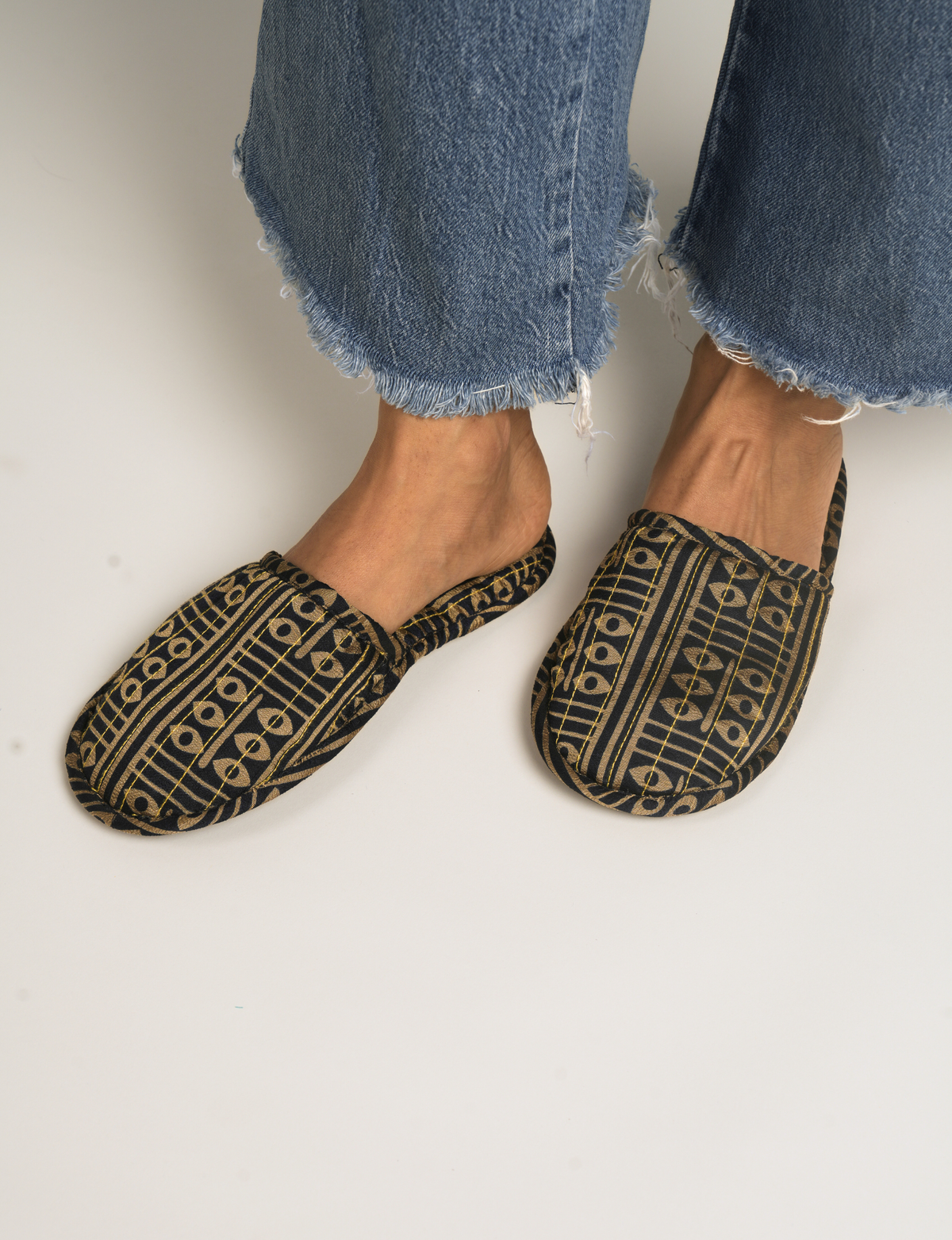 Black and gold patterned slip-on slippers worn with frayed hem blue jeans, front view of feet and ankles
