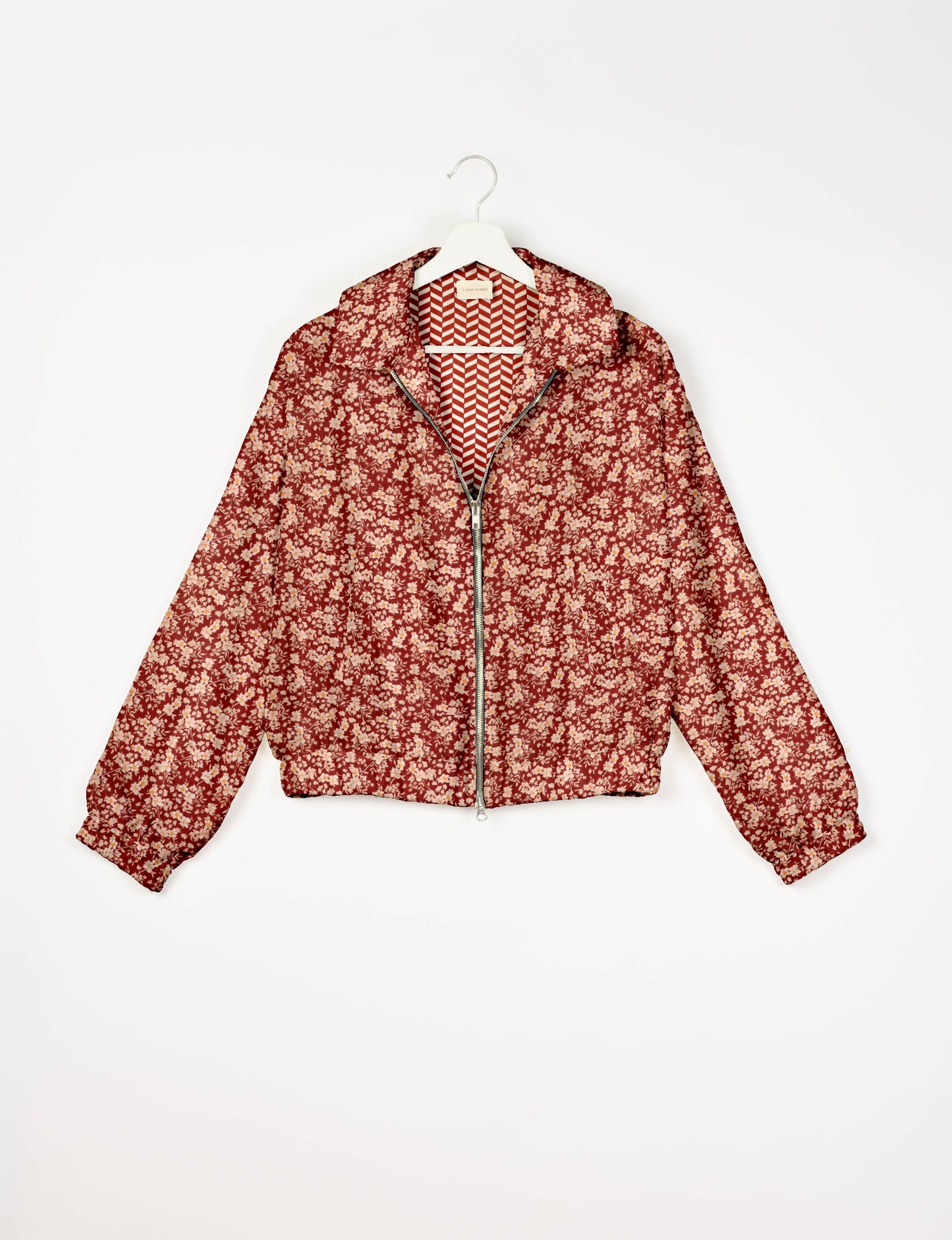 Red floral bomber jacket with front zipper, collar, elastic cuffs, and chevron patterned inner lining, hanging on white