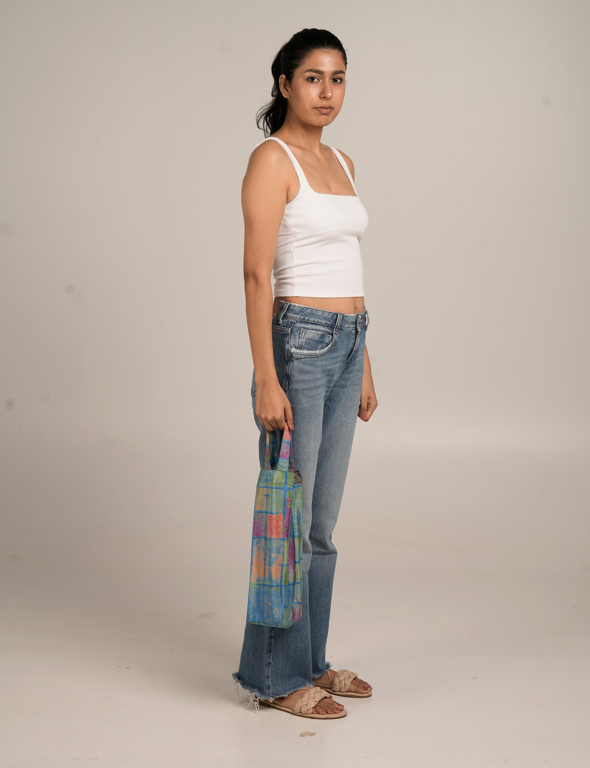 Model holding a blue ethnic-patterned wine bag with multicolor square designs, standing in side view against plain background