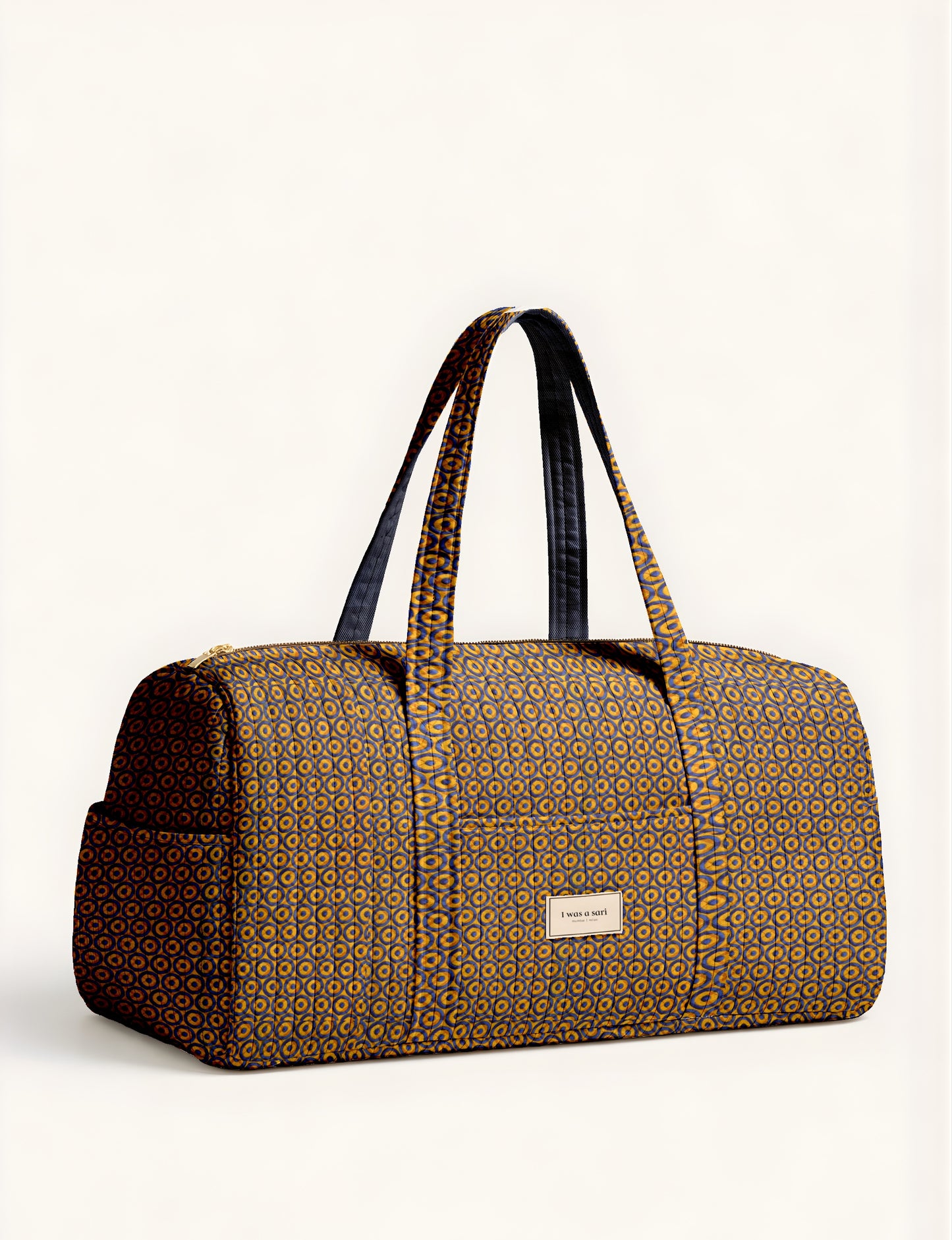 Quilted barrel bag with yellow and dark blue geometric circular pattern, dual handles, front pocket, and "I was a sari"