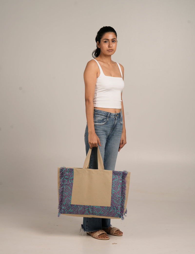 Woman wearing white tank top and blue jeans holding a beige carry-all bag with pink and teal paisley fringe trim