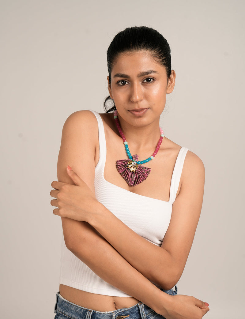 Model wearing a pleated necklace with blue, pink, and white beads and a purple pleated fan-shaped pendant with gold accents
