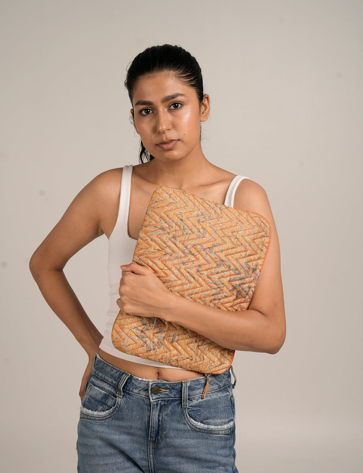 Woman wearing white tank top and blue jeans holding a quilted laptop sleeve with orange paisley pattern in front view
