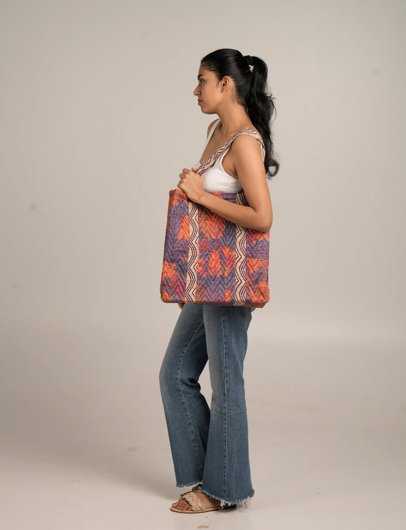 Woman wearing a red and purple quilted tote bag with tropical pattern, paired with white tank top, blue jeans, and beige sandals