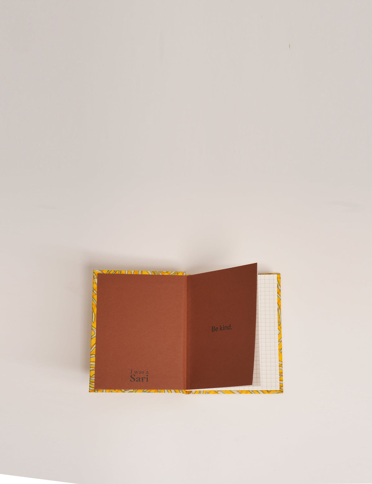 A6 hard cover notebook with yellow abstract patterned cover, open to brown inside cover with "Be kind." and "I was a Sari" text