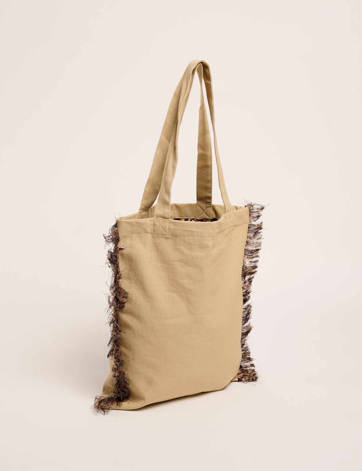 Beige fabric shopper bag with long handles and black fringe detailing along the sides, shown hanging upright
